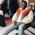 Sean “Diddy” Combs sitting in a chair, wearing sunglasses, a white and orange jacket, and blue jeans.