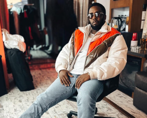 Sean “Diddy” Combs sitting in a chair, wearing sunglasses, a white and orange jacket, and blue jeans.