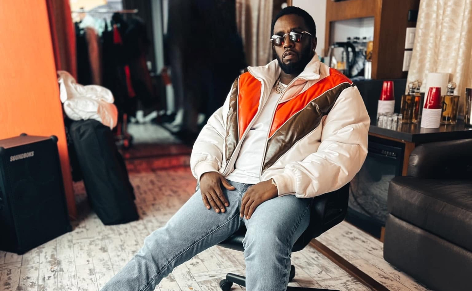 Sean “Diddy” Combs sitting in a chair, wearing sunglasses, a white and orange jacket, and blue jeans.
