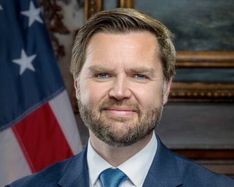 Official portrait of US Vice President JD Vance wearing a blue suit and tie, with an American flag behind him.