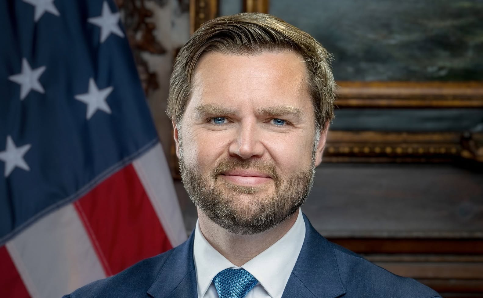 Official portrait of US Vice President JD Vance wearing a blue suit and tie, with an American flag behind him.