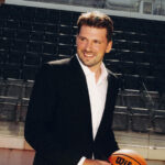 Luka Dončić wearing a black suit and white shirt, smiling while holding a basketball inside an arena.