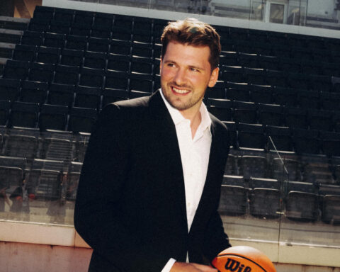 Luka Dončić wearing a black suit and white shirt, smiling while holding a basketball inside an arena.