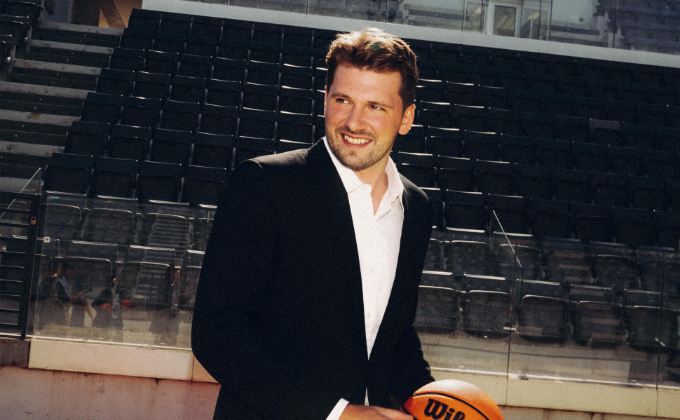 Luka Dončić wearing a black suit and white shirt, smiling while holding a basketball inside an arena.