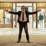 My Cousin Vlad wearing a dark suit, tie and sunglasses, standing with his arms outstretched outside a cinema and theater.