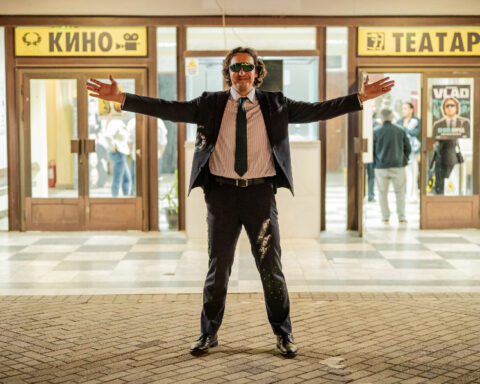 My Cousin Vlad wearing a dark suit, tie and sunglasses, standing with his arms outstretched outside a cinema and theater.