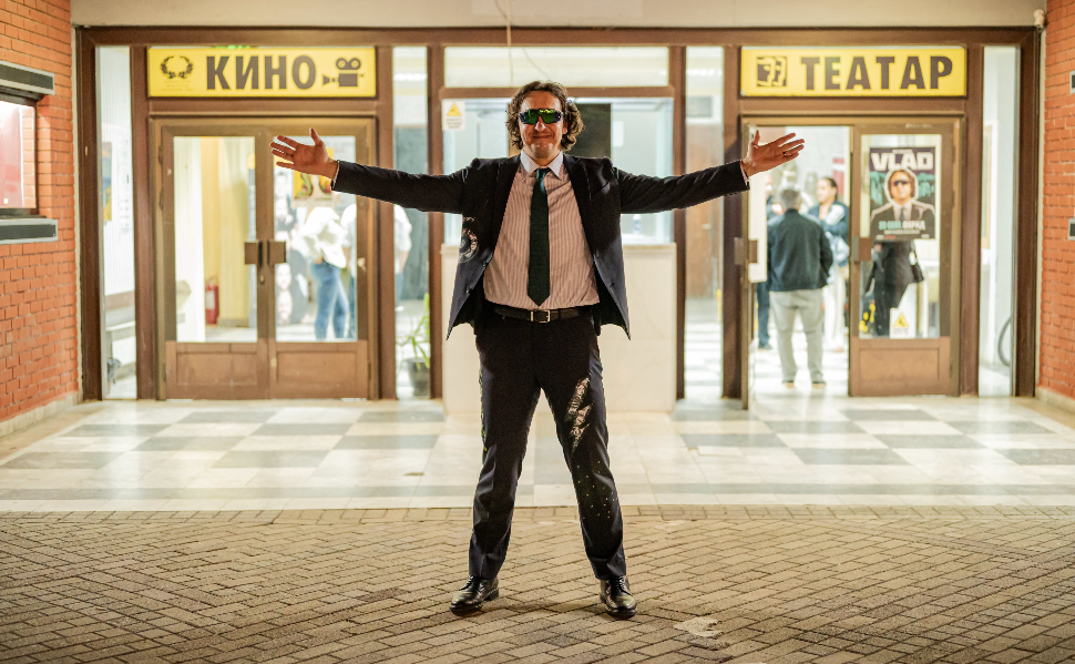 My Cousin Vlad wearing a dark suit, tie and sunglasses, standing with his arms outstretched outside a cinema and theater.
