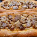 Two freshly baked Macedonian pastrmajlija flatbreads topped with roasted pork pieces and onions on a baking tray.