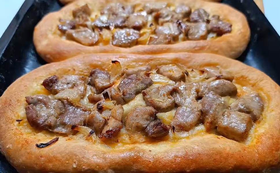 Two freshly baked Macedonian pastrmajlija flatbreads topped with roasted pork pieces and onions on a baking tray.