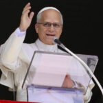 Pope Leo XIV wearing white papal robes, speaking into a microphone and raising his hand.