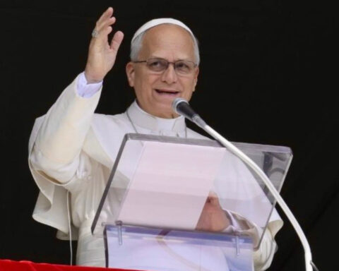 Pope Leo XIV wearing white papal robes, speaking into a microphone and raising his hand.