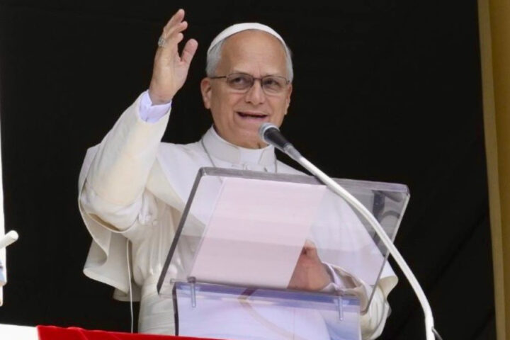 Pope Leo XIV wearing white papal robes, speaking into a microphone and raising his hand.