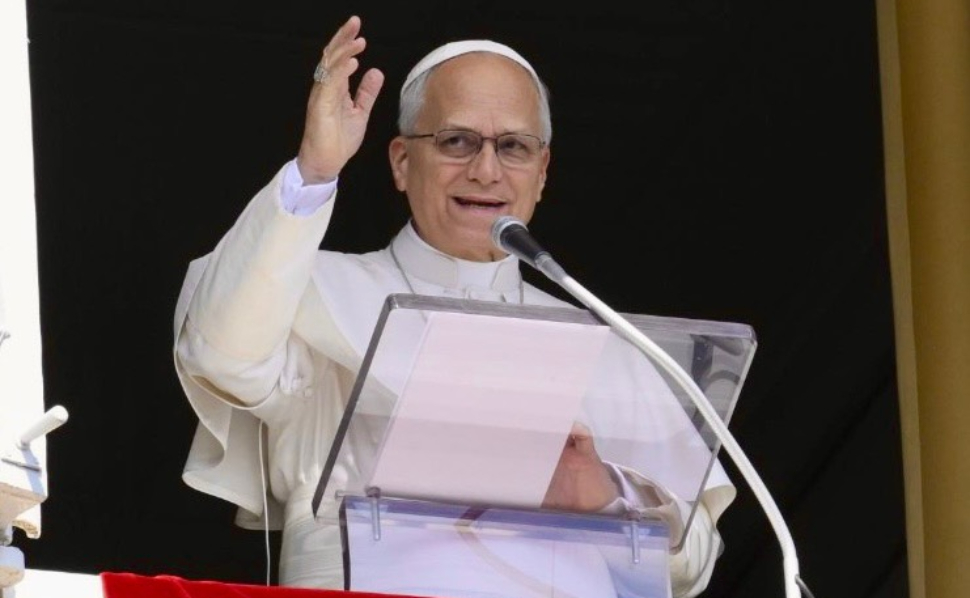 Pope Leo XIV wearing white papal robes, speaking into a microphone and raising his hand.