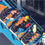 Passengers seated on a roller coaster while staff in high-visibility vests stand alongside the ride.