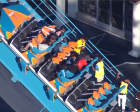Passengers seated on a roller coaster while staff in high-visibility vests stand alongside the ride.