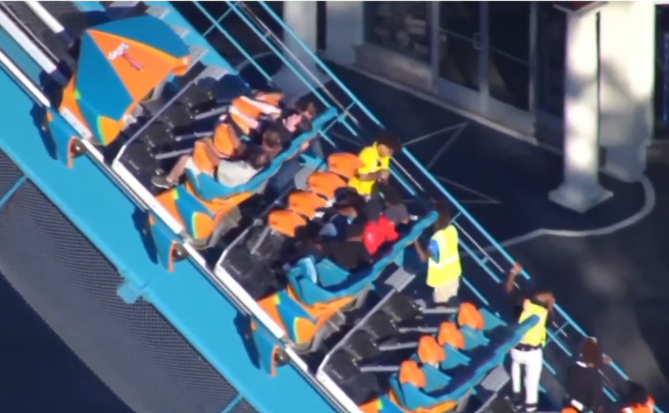 Passengers seated on a roller coaster while staff in high-visibility vests stand alongside the ride.