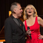 Croatian singer Tajči Cameron in a red dress holding a microphone beside jazz pianist John di Martino on stage.