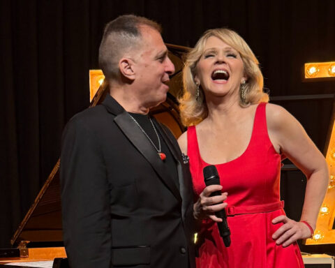 Croatian singer Tajči Cameron in a red dress holding a microphone beside jazz pianist John di Martino on stage.