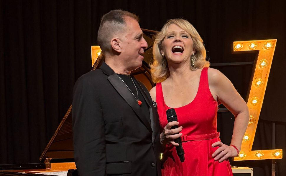 Croatian singer Tajči Cameron in a red dress holding a microphone beside jazz pianist John di Martino on stage.