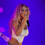 Serbian singer Tea Tairovic performing on stage with a microphone, wearing a white top under purple concert lighting.