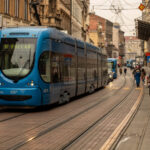 Blue tram traveling through a street in central Zagreb, Croatia, with pedestrians walking along the sidewalks.