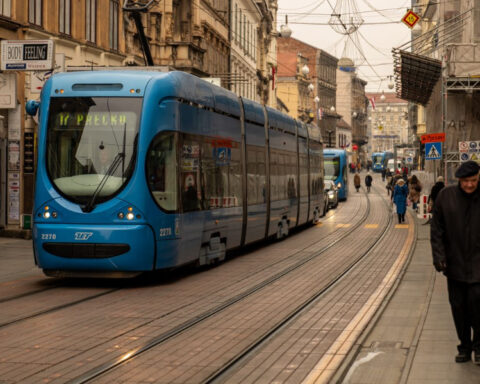 Blue tram traveling through a street in central Zagreb, Croatia, with pedestrians walking along the sidewalks.