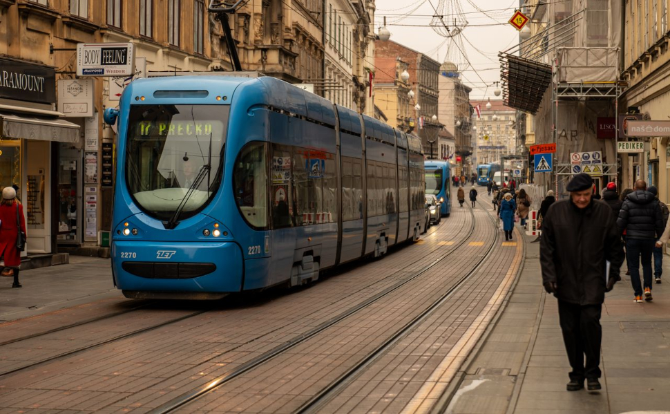 Blue tram traveling through a street in central Zagreb, Croatia, with pedestrians walking along the sidewalks.