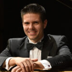 Croatian pianist and composer Bruno Vlahek smiling beside a grand piano.