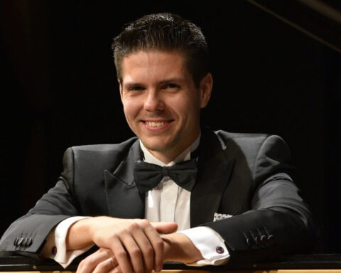 Croatian pianist and composer Bruno Vlahek smiling beside a grand piano.