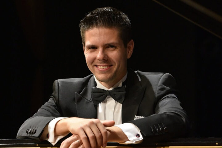 Croatian pianist and composer Bruno Vlahek smiling beside a grand piano.