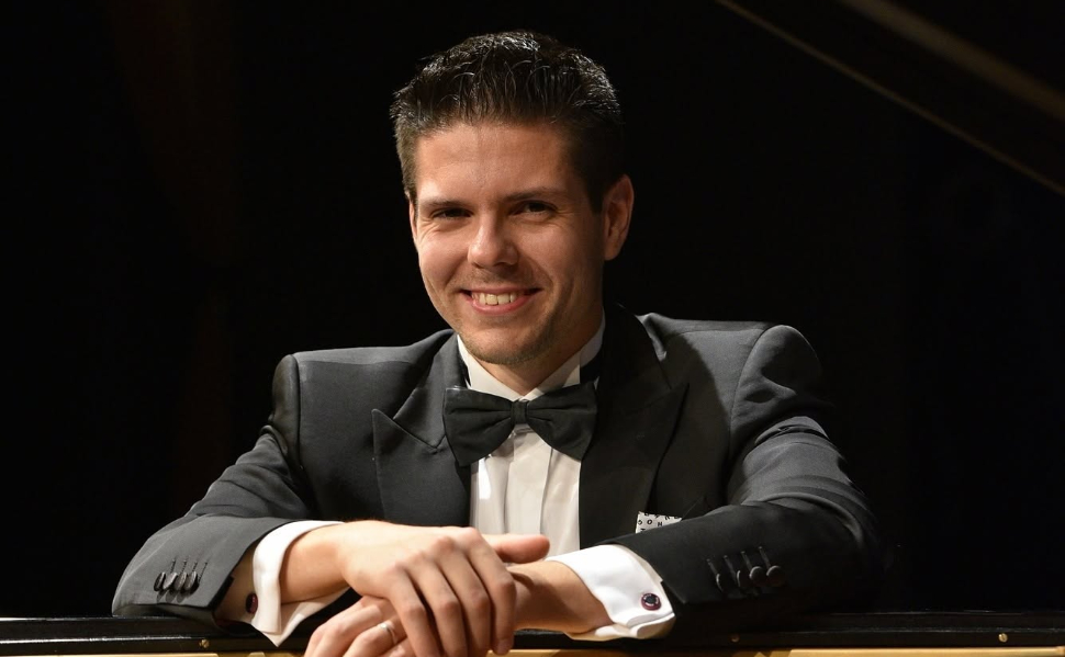 Croatian pianist and composer Bruno Vlahek smiling beside a grand piano.