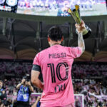 Lionel Messi wearing Inter Miami's pink number 10 jersey and raising a trophy in front of supporters at a stadium.