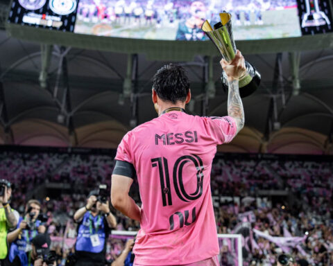 Lionel Messi wearing Inter Miami's pink number 10 jersey and raising a trophy in front of supporters at a stadium.