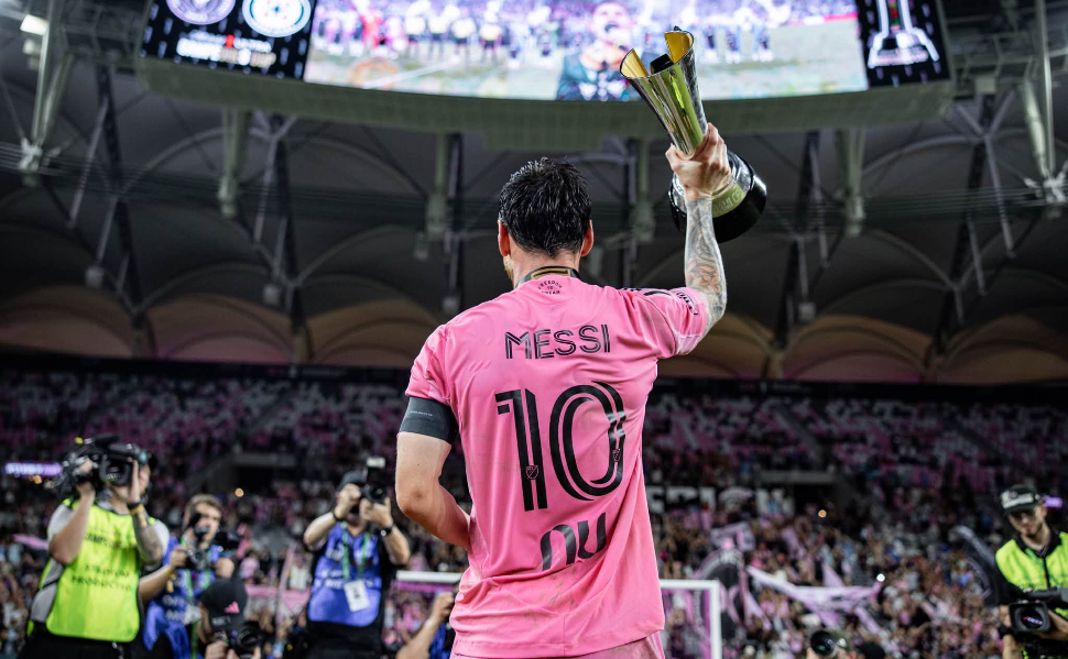 Lionel Messi wearing Inter Miami's pink number 10 jersey and raising a trophy in front of supporters at a stadium.
