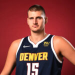 Serbian NBA star Nikola Jokić wearing his Denver Nuggets No. 15 jersey and holding a basketball against a red background.
