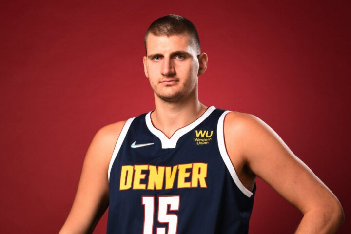Serbian NBA star Nikola Jokić wearing his Denver Nuggets No. 15 jersey and holding a basketball against a red background.