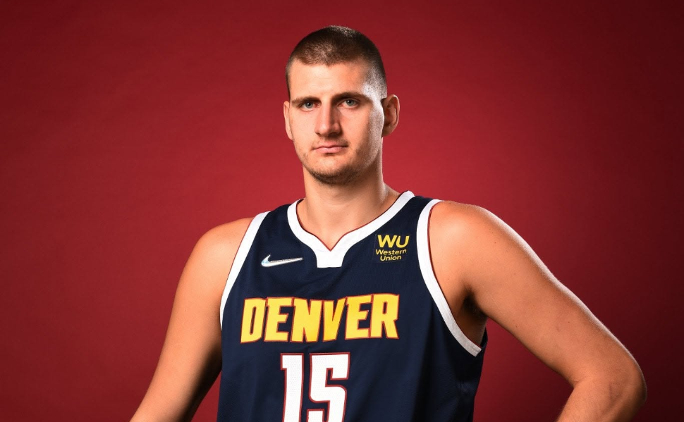 Serbian NBA star Nikola Jokić wearing his Denver Nuggets No. 15 jersey and holding a basketball against a red background.